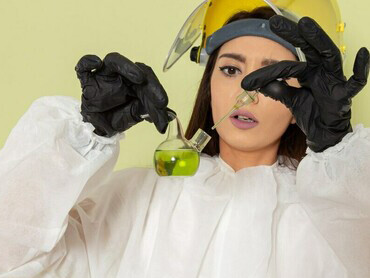 A female chemist in a protective suit holding a green solution on a green surface, cautiously smelling it as part of her experiment