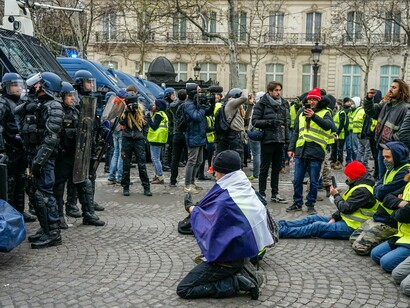 A protester kneeling on the pavement as police watch the crowd, symbolising the contest between authority and dissent