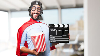 A man dressed in Superhero attire holds popcorn in one hand and a black movie production clapper board 