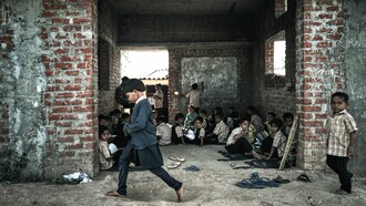 A group of children in school uniforms sit on the dusty floor of an unfinished brick building, a powerful reminder of the urgent need for global efforts to ensure equal opportunities for all