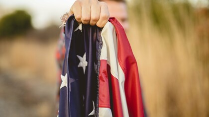 A person holds a tightly folded American flag in their hand, symbolizing national identity and the complex questions surrounding what truly makes a nation great
