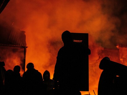 A crowd of silhouetted figures stands in a dark field, positioned behind police barriers under the night sky