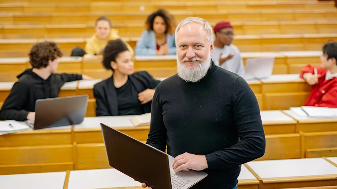 A professor wearing a black turtleneck sweater holds a silver MacBook while teaching in a classroom, encouraging active learning at the university
