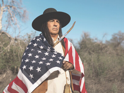 A Native American man standing in the desert with an American flag reflects the complex relationship between American individualism and indigenous identity