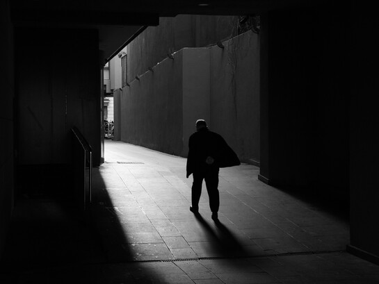 Una figura solitaria avanza por un pasaje oscuro hacia una zona iluminada, dejando una larga sombra sobre el suelo
