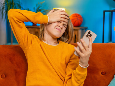 Upset teenage girl sitting on a sofa indoors, reacting to losing an online game and dealing with notification overload on her smartphone