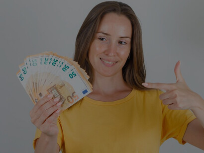 A young woman in a bright yellow t-shirt joyfully points at a stack of euro banknotes, a smile lighting up her face