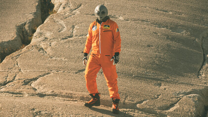 An alien humanoid figure in an orange jumpsuit looks at the camera