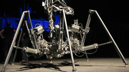 O exoesqueleto de seis pernas de Stelarc. Ao longo de sua carreira, Stelarc realizou mais de 30 performances, sempre explorando a plasticidade do corpo e o hibridismo com a tecnologia. Uma das mais conhecidas é o “Terceiro Braço Robótico”, uma prótese mecânica que se integrava ao seu sistema muscular e se movia através de comandos elétricos, transformando sua anatomia em uma plataforma expandida. Em outras performances, suspendeu seu próprio corpo por ganchos perfurados na pele, numa alusão aos rituais de suspensão corporal, mas também como metáfora extrema para a resistência física e a redefinição dos limites do corpo humano. Talvez uma das imagens mais impactantes de sua trajetória seja a do exoesqueleto de seis pernas, uma enorme máquina que ele controlava para se locomover, assumindo a forma de uma criatura híbrida entre humano e aranha