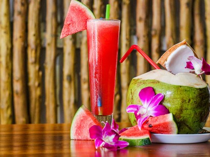Coco fresco decorado com orquídeas cor-de-rosa divide a mesa com uma bebida de melancia, criando uma composição marcada por cores e sabores tropicais. A combinação reforça o caráter sensorial de uma viagem pelo litoral nordestino, onde o mar, a gastronomia e a paisagem fazem parte de uma mesma experiência