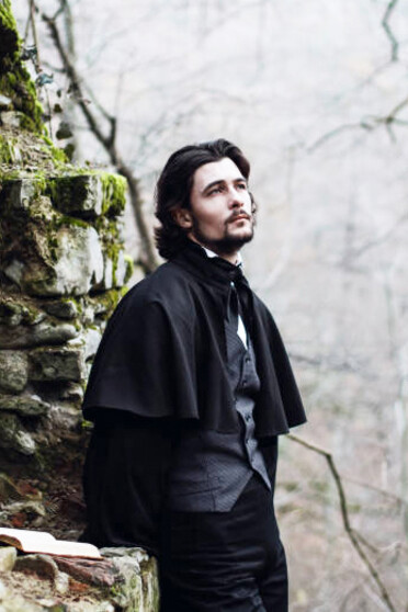 A young man wearing a Victorian-era suit stands against a stone wall