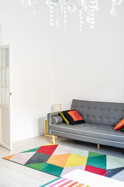 Um sofá cinza se destaca diante da parede clara, enquanto um tapete colorido e elementos decorativos acrescentam personalidade à sala. Como observa o texto, móveis, cores e objetos também podem revelar a relação de seus moradores com a estética e com a imagem que desejam projetar