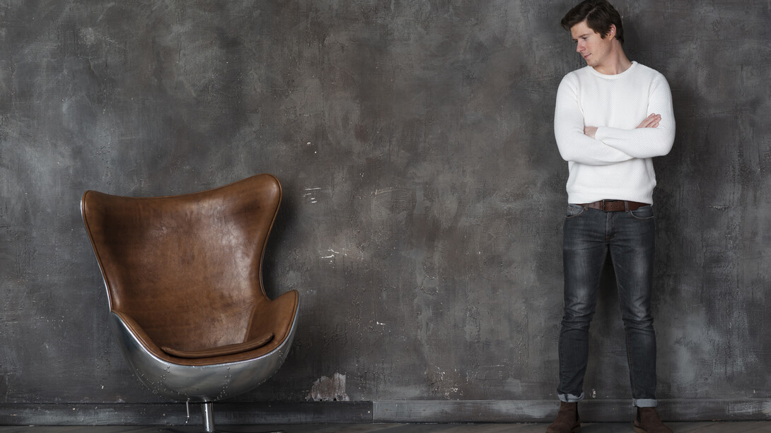 A man stands facing an empty leather armchair, appearing reflective and contemplative