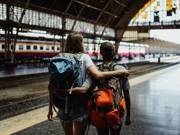 Vistas de costas, duas mulheres caminham juntas pela estação, cada uma levando sua mochila. Ter referências de companhia e segurança durante o trajeto pode tornar mais confortável a experiência de deixar para trás o ambiente familiar
