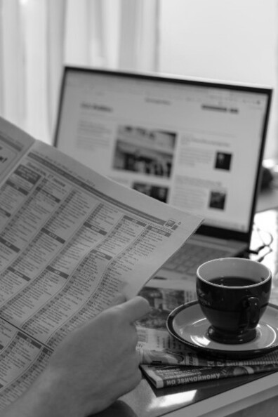 While a man reads a newspaper filled with stories about American politicians, a laptop on the desk beside him displays similar political news online, capturing the blend of traditional and digital media