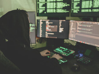 A hacker working across several monitors in a dark room, symbolizing cyber warfare and modern cyber threats