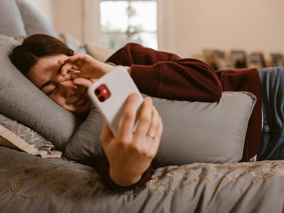 A woman lies in bed using her smartphone, smiling softly as she talks to her boyfriend from afar