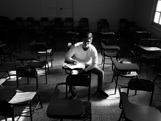 Un estudiante escribe a mano en el interior de un aula prácticamente vacía, rodeado de pupitres desocupados y sin ningún compañero a la vista