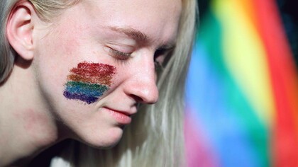 A trans woman with rainbow glitter on her cheek and the rainbow flag in the background