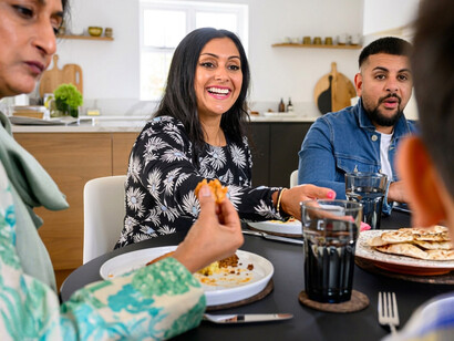 Three generations of a Pakistani family gather around the table for dinner, sharing food and conversation while a woman cooks nearby