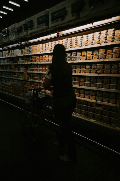A woman with a shopping cart stands in a softly lit supermarket aisle, surrounded by rows of neatly stocked products