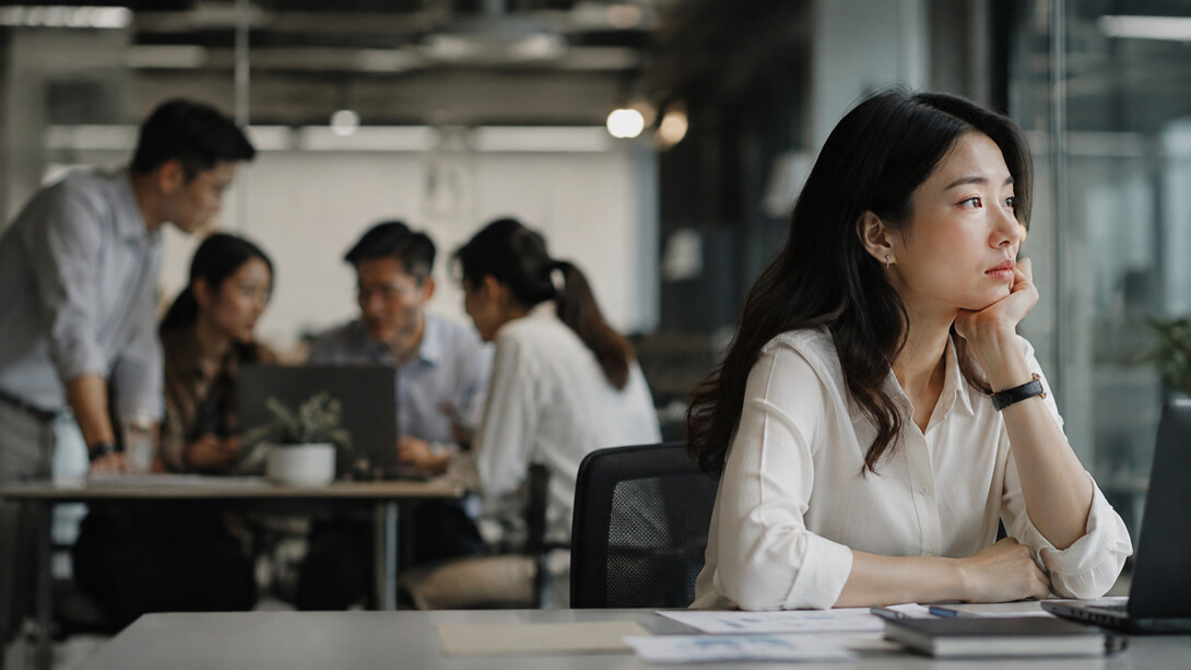 A woman sitting apart from her coworkers in a busy office, representing the emotional distance that can exist within a workplace