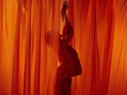 Em uma pose marcada pelo movimento, o bailarino se deixa envolver pelas cortinas em tons de vermelho e laranja. O fogo continua aceso quando a criação deixa de ser apenas uma atividade e passa a fazer parte da própria maneira de existir. Talvez seja isso que significa continuar vivo: mudar a forma da dança sem perder a chama que a faz nascer