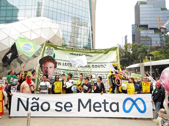 Manifestantes se reúnem em São Paulo diante da sede da Meta, empresa responsável por plataformas como Facebook, Instagram e WhatsApp, para protestar contra a possível interferência das redes sociais no processo eleitoral brasileiro. A faixa central resume a reivindicação: “Não se Meta”, em um jogo de palavras que transforma o nome da companhia em uma denúncia política. Entre bandeiras, cartazes e imagens de figuras públicas, outras mensagens pedem: “Tira a mão da eleição”. A manifestação expressa a preocupação com o poder das grandes plataformas de influenciar o debate público, determinar a visibilidade de discursos e interferir na maneira como informações e opiniões circulam. Em uma sociedade na qual a disputa política também se trava por meio de algoritmos, memes e campanhas digitais, a fotografia evoca uma questão central: quem controla os mecanismos que ajudam a formar a opinião pública e até que ponto esse poder pode afetar a democracia? A fotografia corresponde ao protesto realizado em 19 de outubro de 2026, diante da sede da Meta, na Avenida Faria Lima, em São Paulo