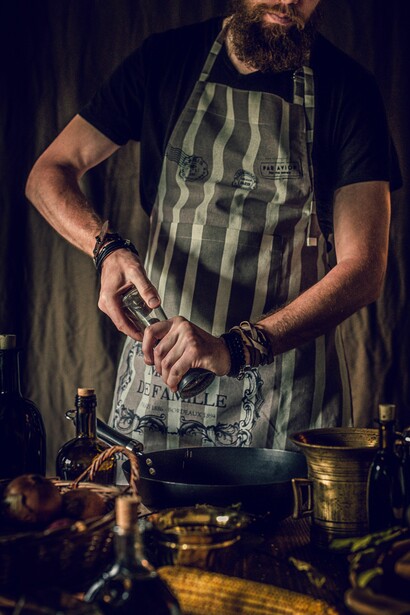 A bearded chef holds a frying pan in a rustic kitchen surrounded by fresh ingredients and traditional cooking utensils