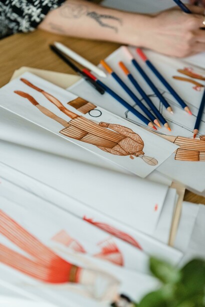 Mesa de trabalho com rascunhos de peças de roupa e um braço desenhando. Entre lápis e papel, surgem propostas que unem inovação, arte e responsabilidade