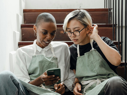 Colleagues wearing aprons sitting on a staircase, scrolling through social media on their smartphones  