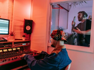Produtor musical acompanha a gravação enquanto o cantor se apresenta do outro lado do vidro do estúdio. A tecnologia integra o processo, mas não substitui a intenção de quem cria