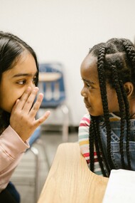 Two girls whispering during a lesson, inside a classroom, at school