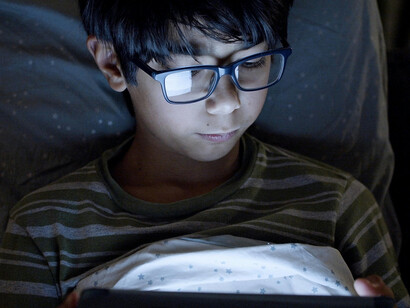 A boy wearing eyeglasses uses a digital tablet at night, focused on the screen