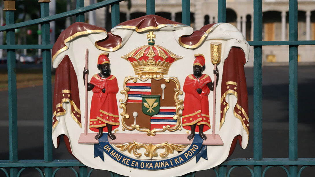 The coat of arms is divided into four quarters. The first and fourth feature eight alternating stripes of white, red, and blue, drawn from the Hawaiian flag and symbolizing the kingdom’s eight inhabited islands