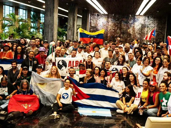 Integrantes da ALBA Movimentos reúnem-se durante a 4ª Assembleia Continental da organização, em Havana, diante das bandeiras de Cuba, Venezuela e Argentina. A imagem traduz a articulação política entre movimentos populares latino-americanos que o artigo apresenta como parte da disputa pela soberania e contra a ingerência imperial na região. A diversidade de movimentos representada na cena traduz o projeto de integração popular e solidariedade internacionalista que está no centro da articulação. A Alba Movimentos (Articulação Continental de Movimentos Sociais e Populares rumo à Alba) é uma rede que une centenas de organizações populares da América Latina e do Caribe — como sindicatos, grupos feministas, movimentos estudantis, camponeses e indígenas. Inspirada na aliança de governos da ALBA-TCP, ela atua de forma autônoma na base da sociedade para promover a integração regional e a resistência anticapitalista