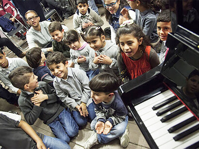 Kids from Africa, Syria, Afghanistan, Pakistan,  set in a semi-circle around their teacher, ph. El Sistema Greece