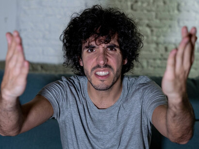 A disappointed young man watches television from his couch, shouting and gesturing angrily at the scree