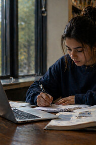 A young woman focused on her assignment, surrounded by textbooks and a laptop, symbolising the combination of traditional and digital education