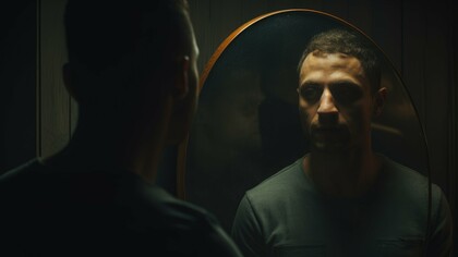 A dimly lit portrait of a man staring into a mirror, his reflection revealing a shadowed, contemplative expression, captures the essence of shadow work—facing the parts of ourselves we often ignore