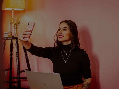 A woman in a café has her laptop open while she's taking a selfie from her phone to help quench that thirst for social media engagement