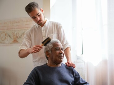 Um homem de camisa branca penteia os cabelos de um homem idoso que veste um suéter azul, junto a uma janela iluminada pela luz natural. O gesto cotidiano revela uma forma de cuidado que se torna especialmente significativa quando a passagem do tempo modifica a autonomia e as necessidades de uma pessoa. Receber ajuda não diminui uma vida: revela a importância dos vínculos que a sustentam