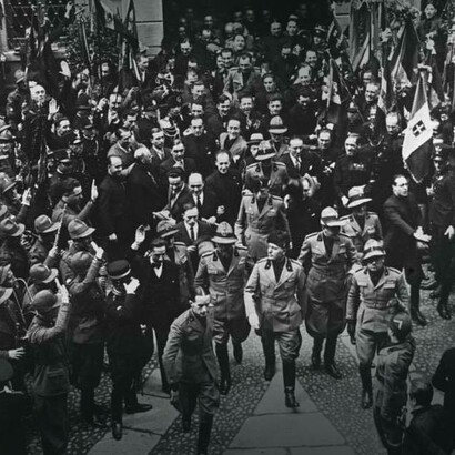 Il discorso politico di Mussolini a Milano. Benito Mussolini, accompagnato dai suoi sostenitori sulla piazza del Duomo di Milano, si dirige verso la tribuna, maggio 1930-1