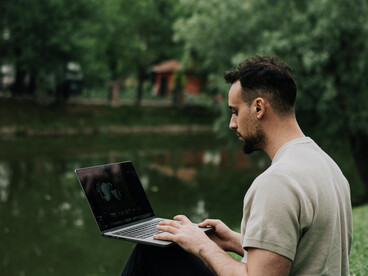 A man relaxes on the grass beside a peaceful pond in a park, working on his laptop while enjoying the natural surroundings