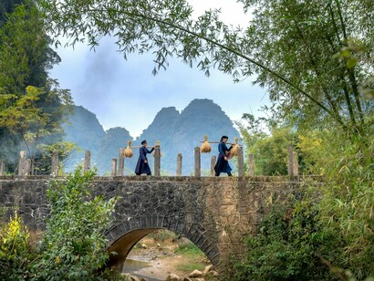 Villagers carrying harvested bundles across an old stone bridge, representing the burdens carried by rural communities