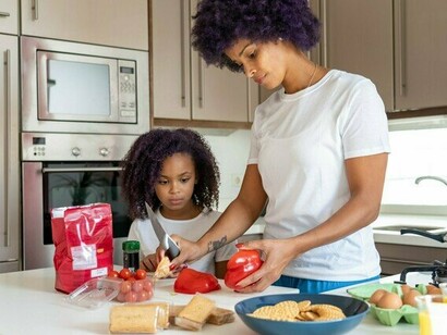 A mother guiding her daughter through meal preparation, representing the passing of practical life skills from one generation to the next