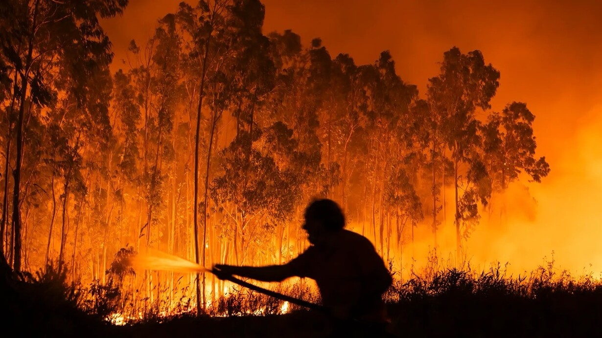 Silhueta de um homem segurando uma mangueira enquanto tenta conter uma floresta completamente tomada pelas chamas. A imagem simboliza a luta desigual contra um problema que se repete todos os anos sem soluções eficazes