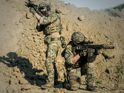 Dois militares vestindo uniformes de camuflagem e portando armas participam de uma atividade tática em uma paisagem desértica. A imagem destaca a preparação e a coordenação entre combatentes, elementos importantes para compreender a dimensão operacional dos conflitos armados contemporâneos