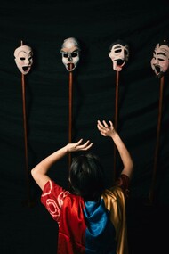 A woman in a robe performs with theater masks against a black background, embodying the tension between identity and performance