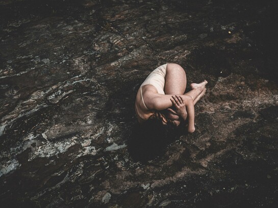Uma mulher permanece encolhida sobre uma superfície áspera, em uma postura que transmite vulnerabilidade e sofrimento. Quando emoções permanecem silenciadas por muito tempo, o corpo pode transformar esse silêncio em sinais que pedem atenção e cuidado. A imagem de uma mulher encolhida sobre uma superfície rochosa busca transmitir vulnerabilidade, sofrimento, recolhimento e tensão emocional, funcionando como uma metáfora visual para o momento em que o corpo passa a expressar aquilo que a mente não conseguiu elaborar. Ansiedade, tensão e outros sintomas físicos podem revelar conflitos emocionais que ainda buscam um caminho de elaboração. Escutar o que o corpo comunica não significa ignorar a medicina, mas reconhecer que algumas dores também carregam histórias que precisam ser compreendidas